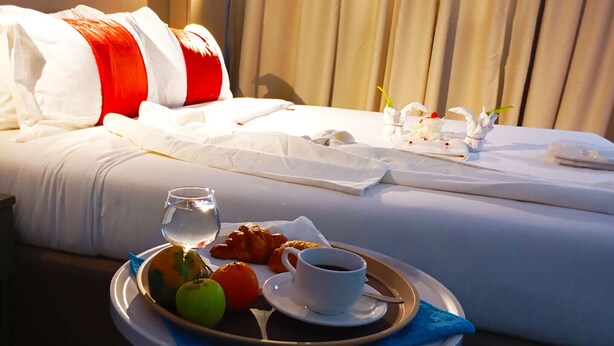 Una cama bien hecha con sábanas blancas y una almohada roja y blanca, una bandeja con frutas y una taza de café, y un pequeño florero con flores sobre la mesita de noche.