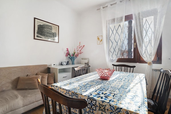 A dining area with a table covered in a patterned tablecloth, a sofa, chairs, a vase with flowers, and a framed picture on the wall.