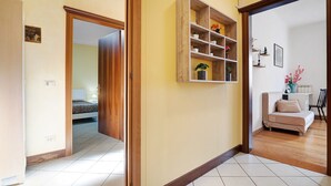 A hallway with a wooden door, a white wall, a wooden shelf with plants, a beige sofa, and a tiled floor.