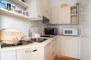 A kitchen with a dishwasher, two microwaves, a toaster, and a wooden cutting board.