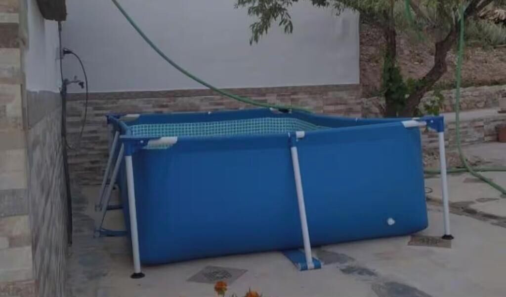 A blue pool with a white frame, set against a stone wall and a tree.