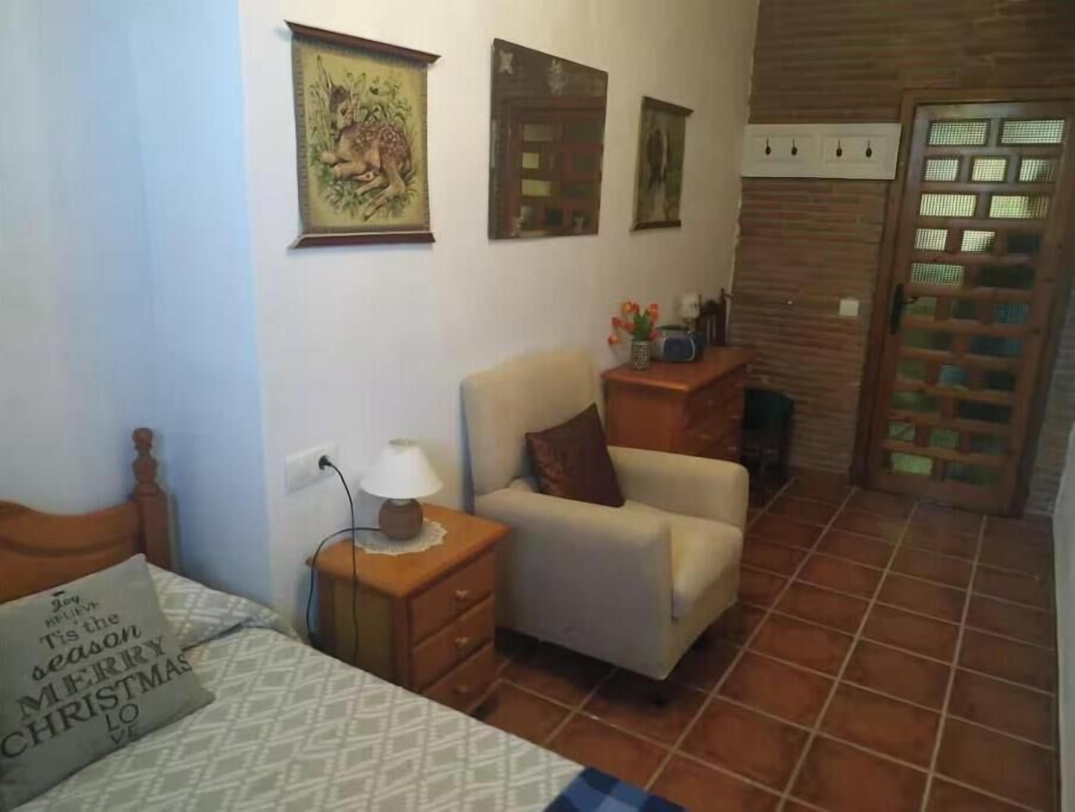 Een slaapkamer met een bed, een nachtkastje met een lamp, een stoel en een dressoir. Er zijn decoraties aan de muur en een deur met een raam.