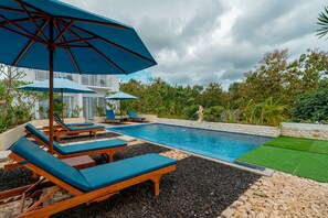 A pool area with lounge chairs and umbrellas, surrounded by a landscaped garden and a building in the background. - Bella Belly Cottage and Villa (Penida Island)