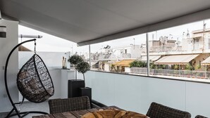 A rooftop terrace with a wooden dining table, wicker chairs, a hanging chair, and a view of the cityscape.