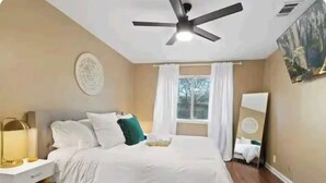 Ein Schlafzimmer mit Bett, Nachttisch, Deckenventilator, einem Fenster mit Vorhängen und einem Gemälde an der Wand.