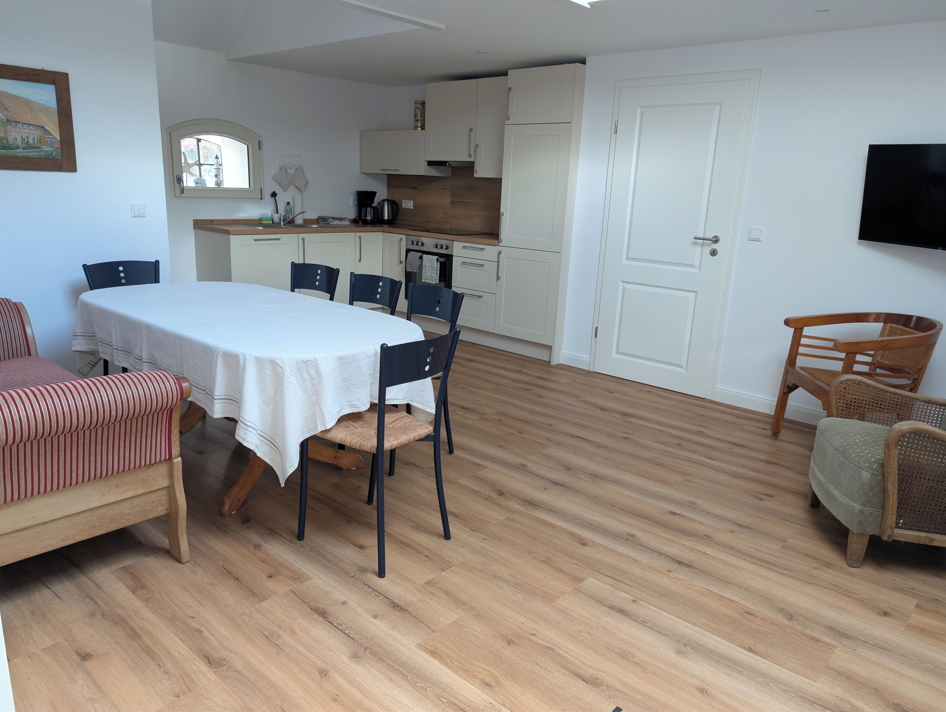A dining area with a table and chairs, a kitchen with white cabinets, a wooden floor, and a door.