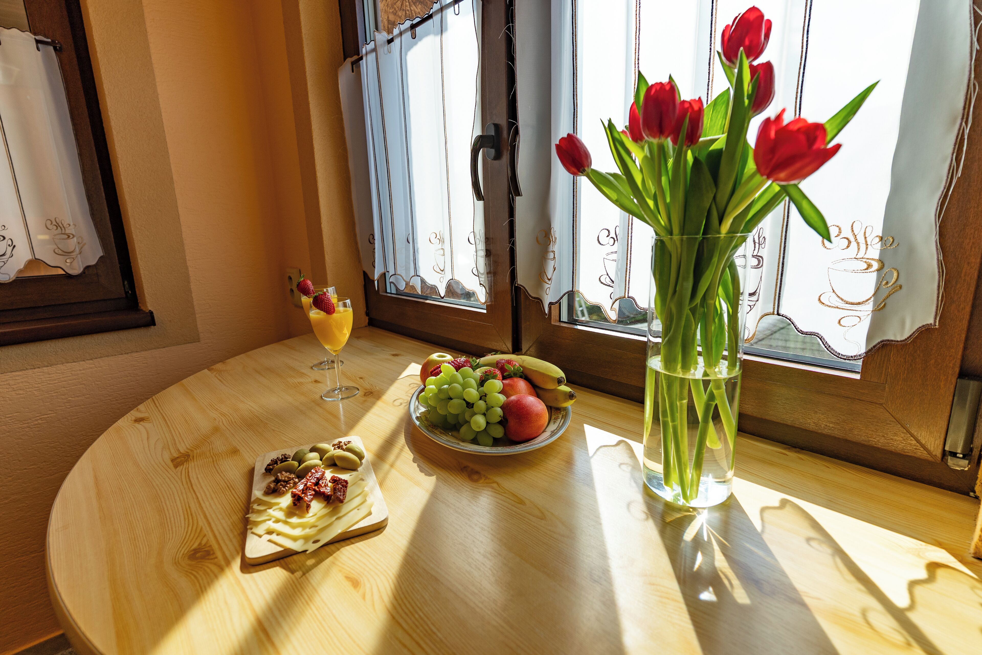 Una mesa de madera con un frutero, un vaso de bebida, un jarrón con flores rojas y un plato con postre.