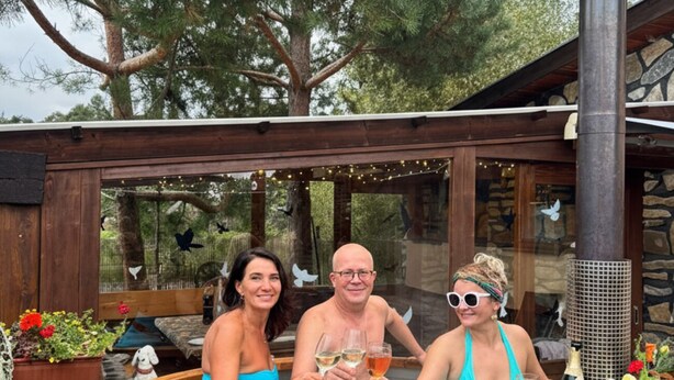 A hot tub with three people toasting with drinks, surrounded by a wooden deck and stone walls, with trees in the background. - Privát Prima (Podturen)