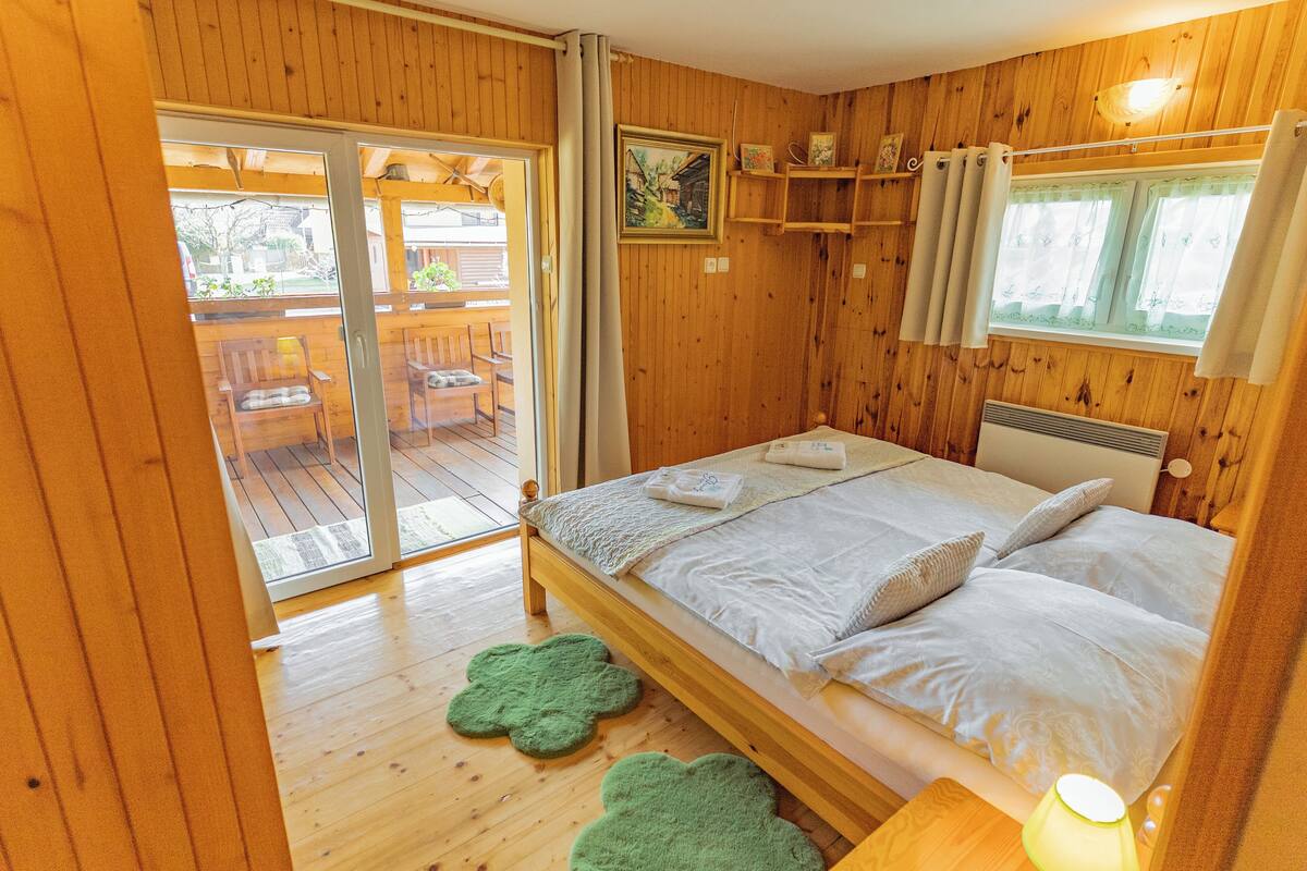 Une chambre à coucher aux murs lambrissés, avec un lit, une porte-fenêtre coulissante donnant sur un espace extérieur et une fenêtre avec des rideaux.