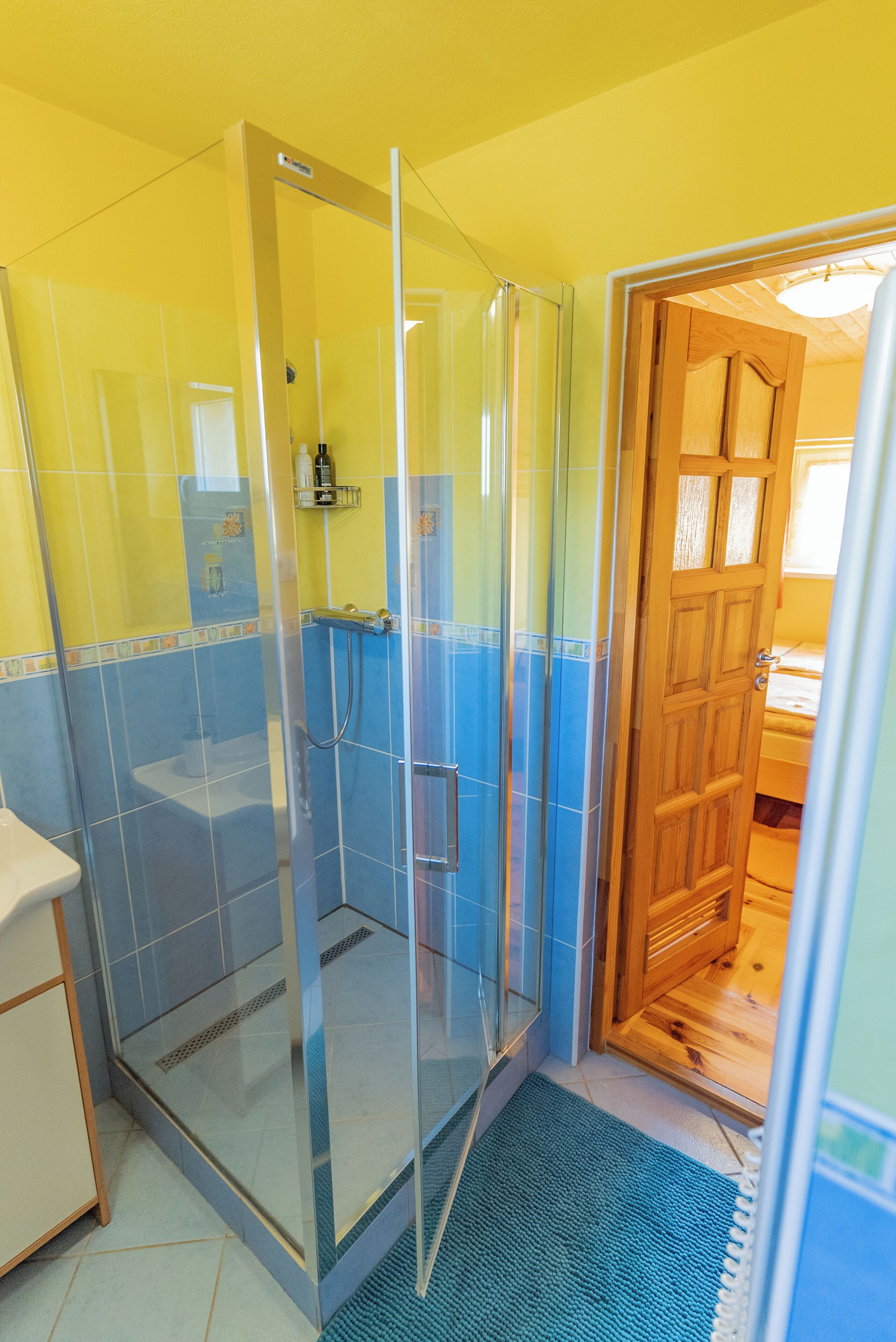Un baño con ducha separada por vidrio, paredes amarillas y una puerta de madera que da a otra habitación.