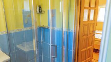 Un baño con ducha separada por vidrio, paredes amarillas y una puerta de madera que da a otra habitación.