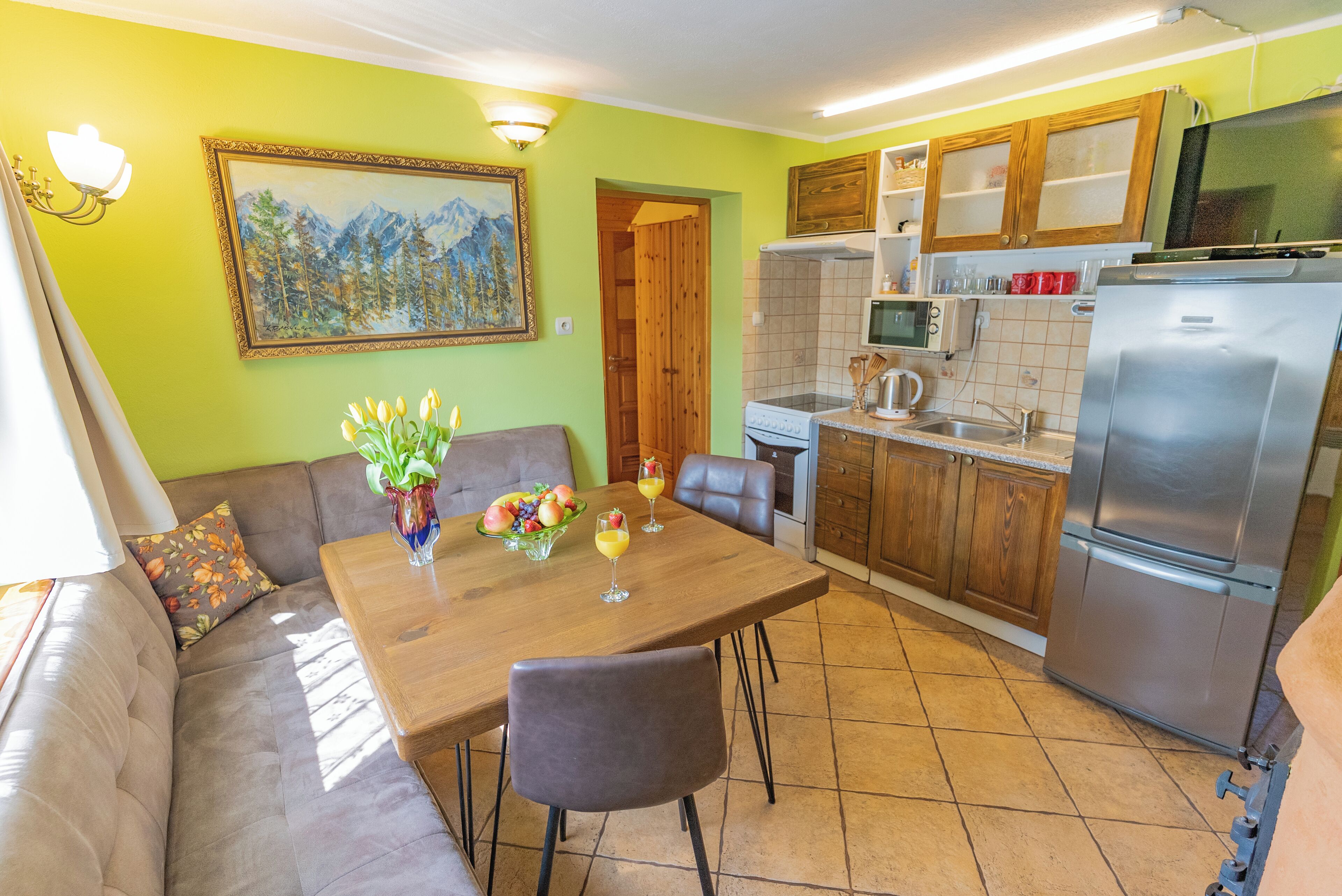 A compact kitchen with a refrigerator, microwave, and stove. There's a dining table with chairs and a sofa with cushions. A painting hangs on the wall, and a vase with flowers is on the table.