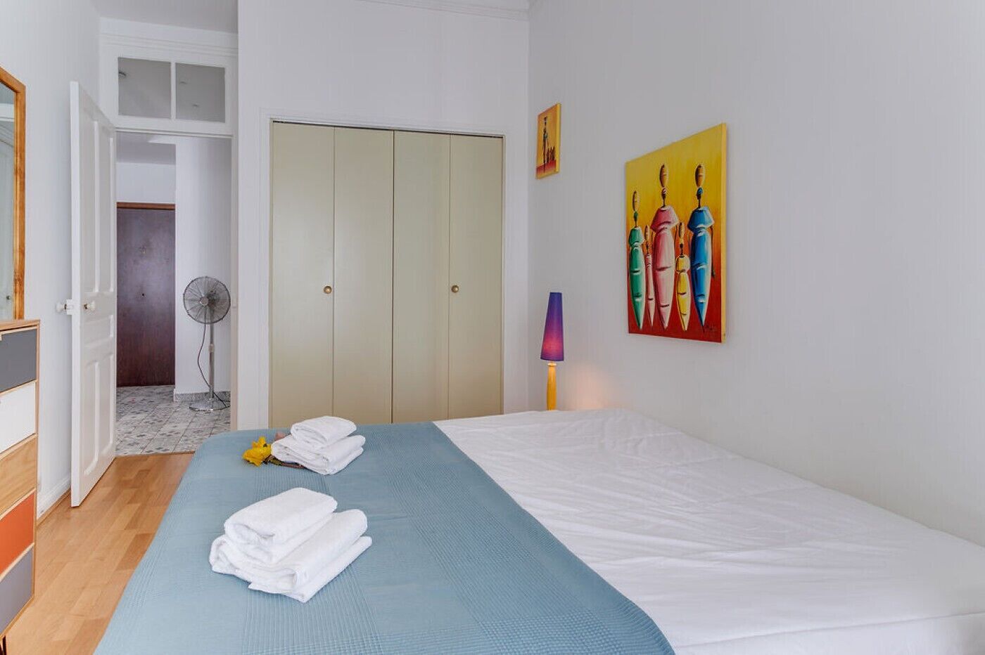 Une chambre d’hôtel avec un lit, un ventilateur, un tableau et une porte donnant sur une salle de bain.