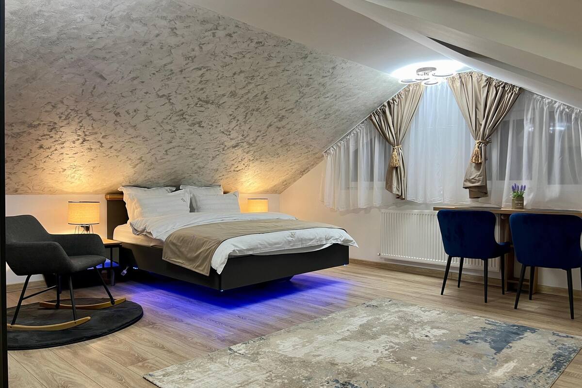 Een slaapkamer met schuine wanden, een bed met hoofdbord, een bureau met twee stoelen, een lamp en een radiator.