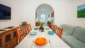 Uma sala de jantar com uma mesa comprida posta para seis pessoas, rodeada por cadeiras, com uma televisão à esquerda e um quadro à direita.