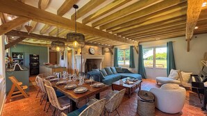 A spacious living room with a large wooden ceiling, a dining area with a long table and wicker chairs, a sofa, and a piano.