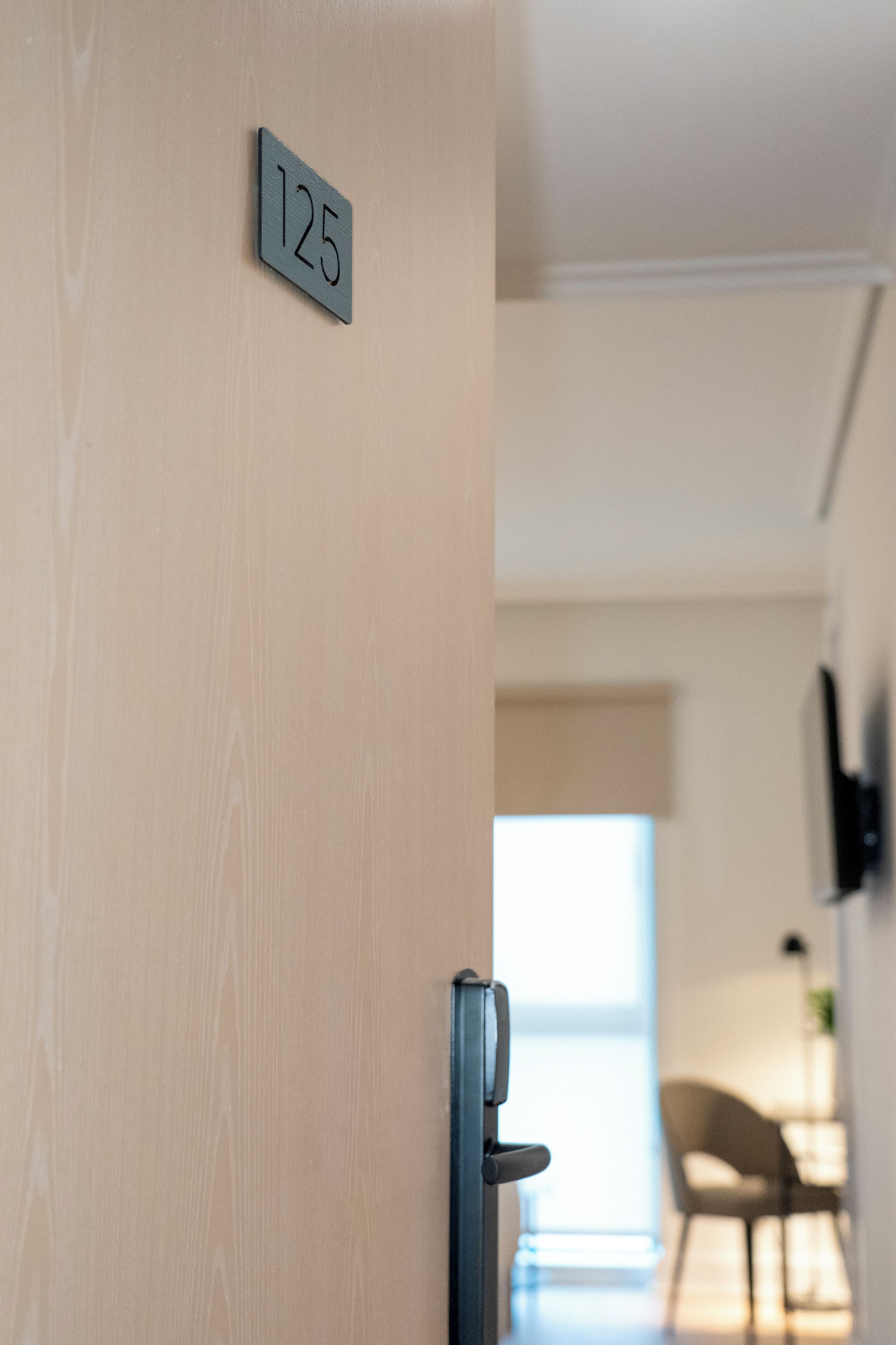 Una puerta con una placa que indica la habitación 125, un picaporte moderno, y una vista parcial de una habitación con una silla y una planta al fondo.