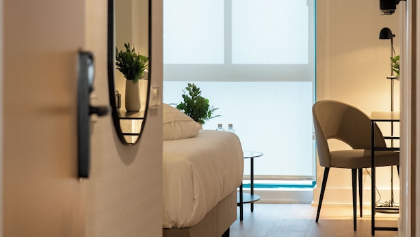 Habitación de hotel con cama, una silla, una mesita, una planta en maceta y un ventanal grande.