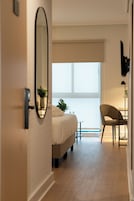 Habitación de hotel con cama, una silla, una mesita, una planta en maceta y un ventanal grande.