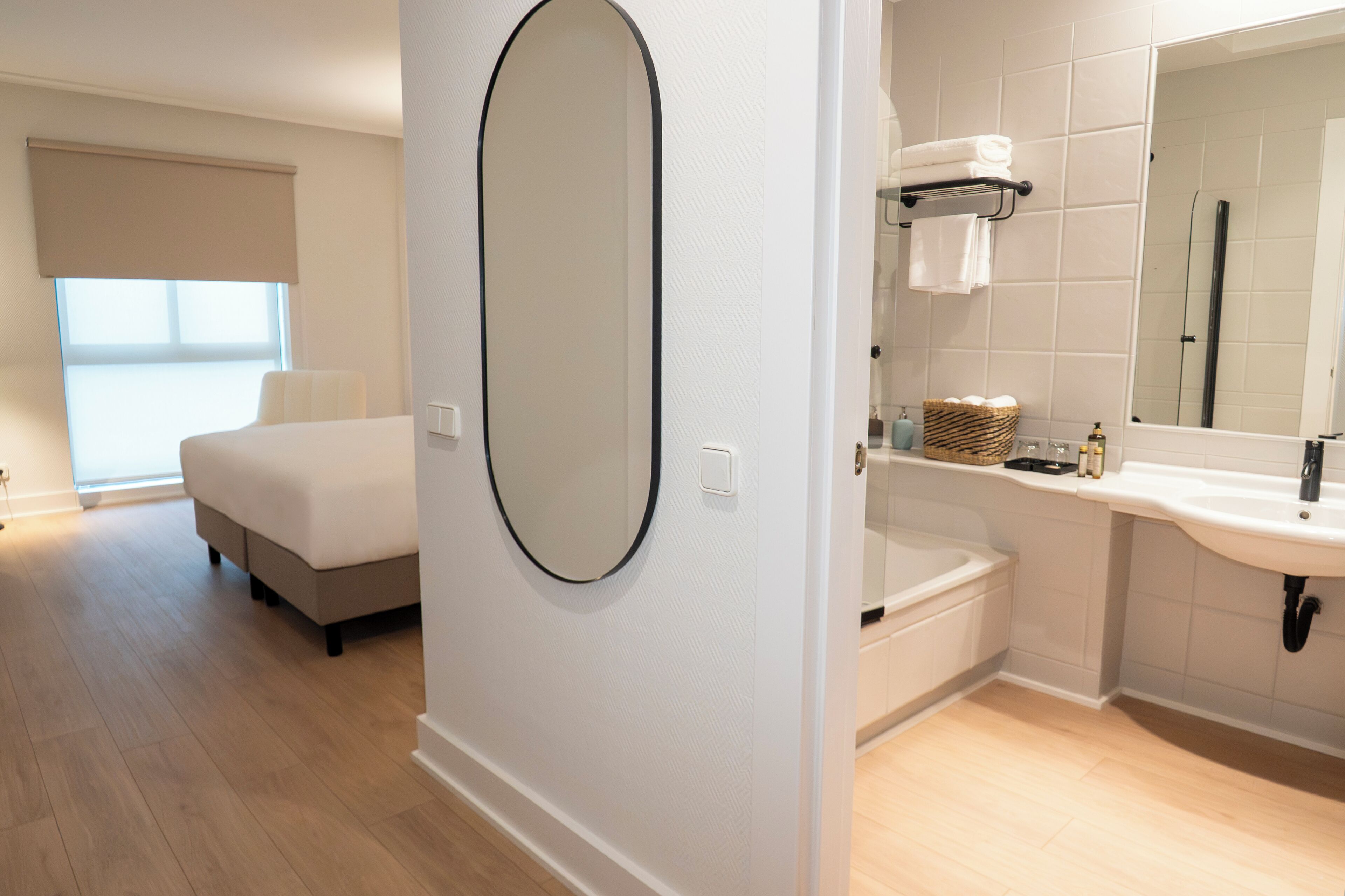 Una habitación de hotel moderna con una cama grande, un espejo redondo, un baño con bañera y un ventanal amplio.