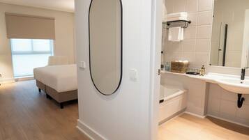 Una habitación de hotel moderna con una cama grande, un espejo redondo, un baño con bañera y un ventanal amplio.