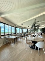 Amplio espacio interior con grandes ventanales, suelo de madera y diversas disposiciones de asientos, incluyendo mesas y sillas.