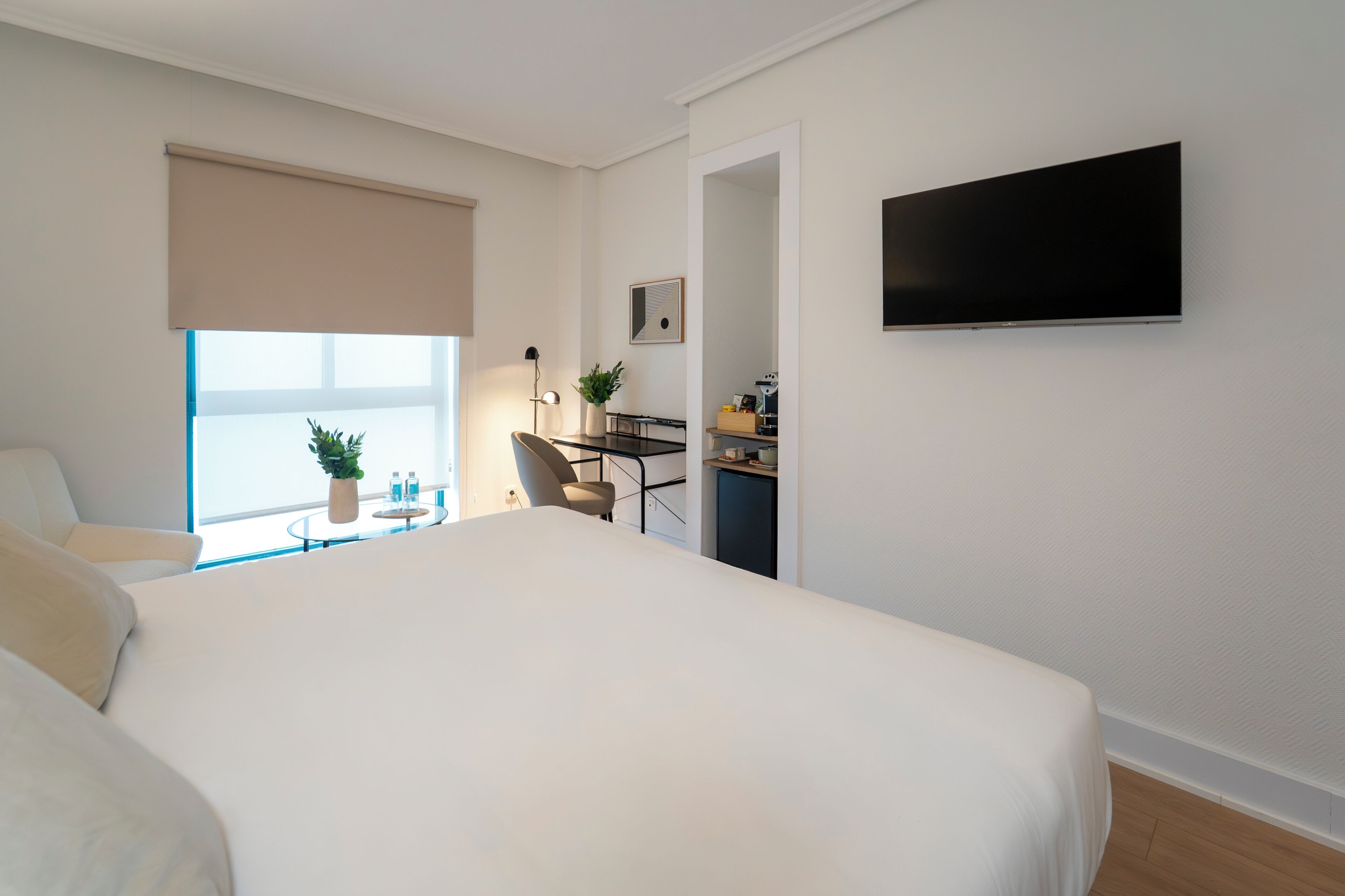 A modern hotel room with a large bed, a desk with a chair, a flat-screen TV mounted on the wall, and a window with a view.