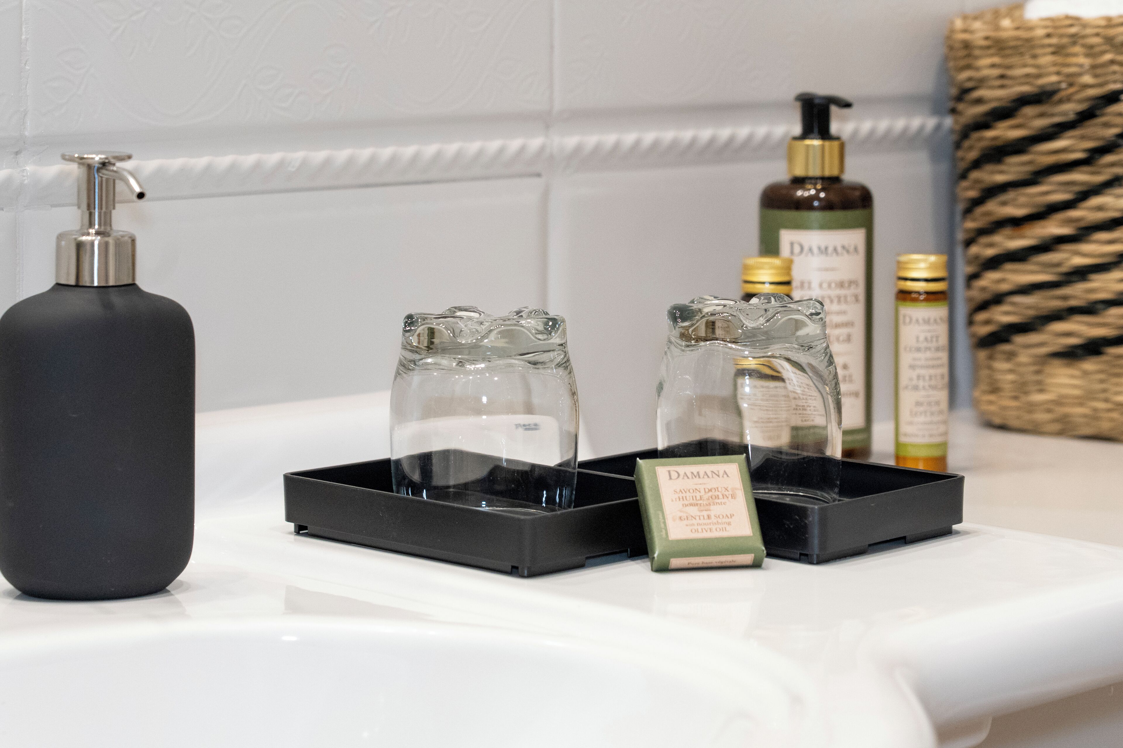 A bathroom counter with a black soap dispenser, two glass soap dishes, and a bottle of Damana soap.