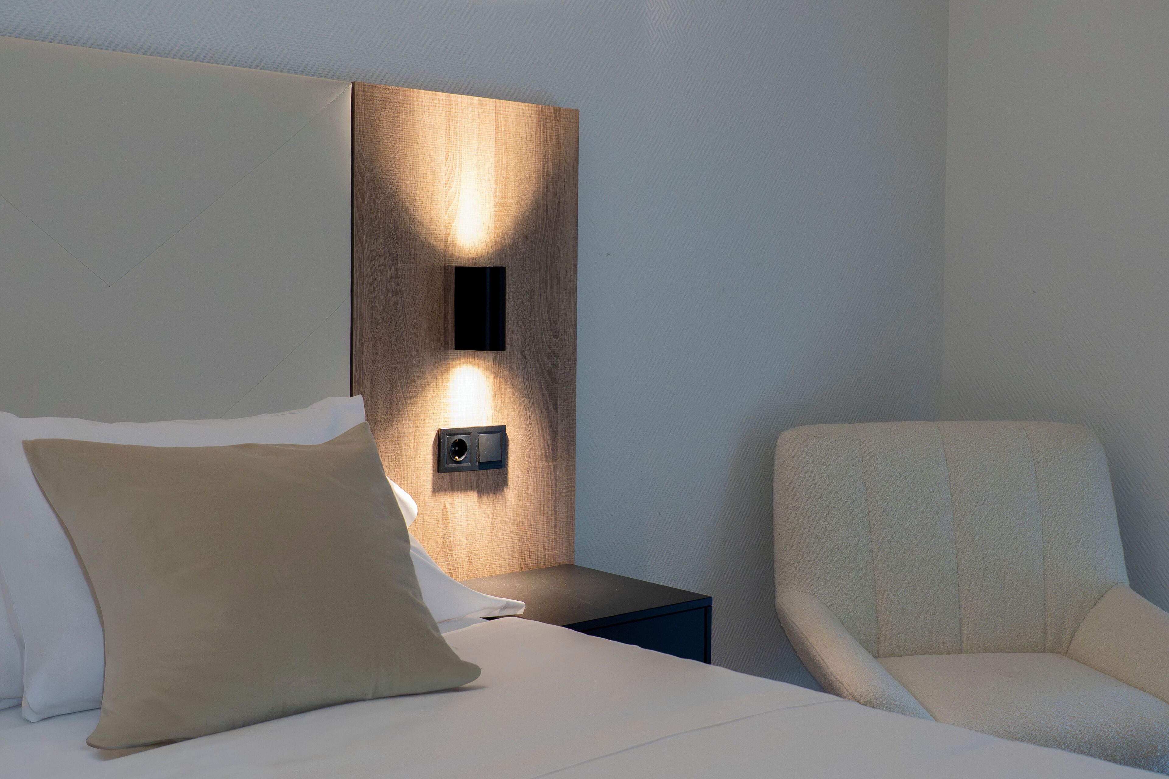 A neatly made bed with pillows, a bedside table with a wall-mounted light, and a chair.