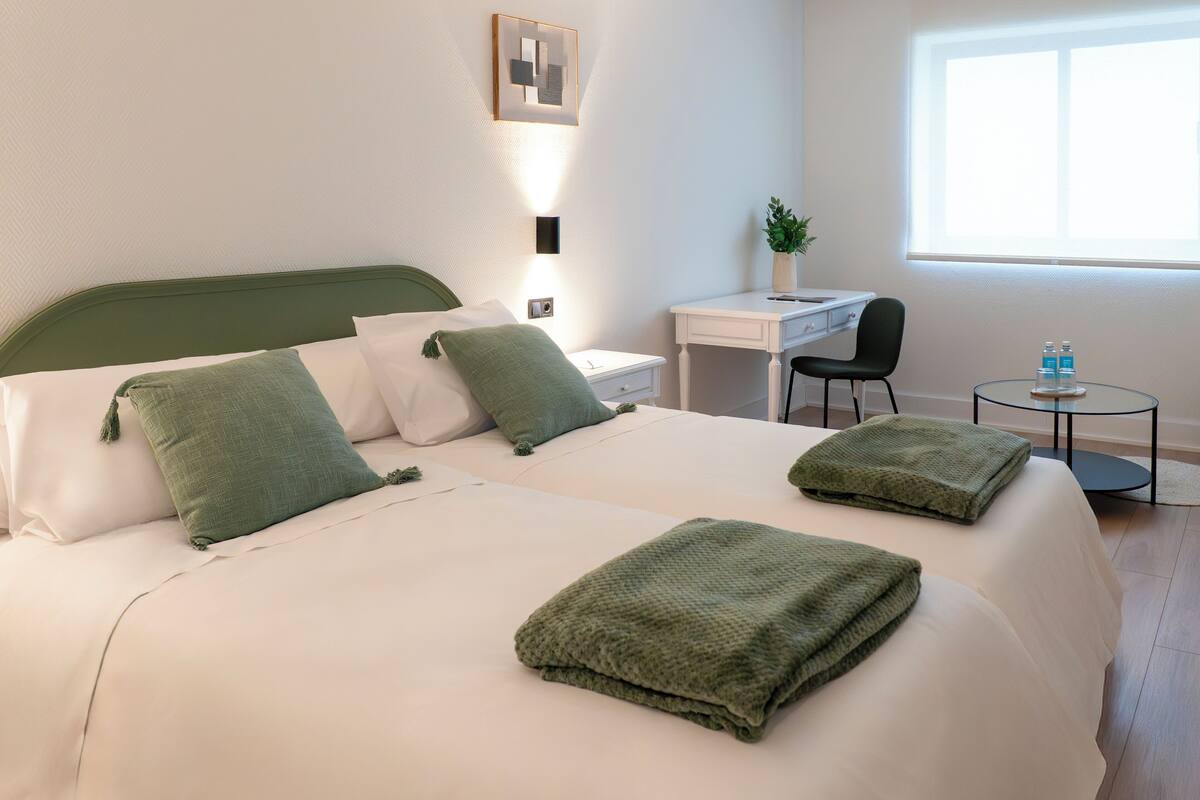 A hotel room with a bed, a desk, a chair, and a small table with bottles.