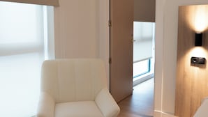 Una habitación moderna con un sillón blanco, una puerta de madera con una lámpara, y vista a un balcón a través de una puerta abierta.