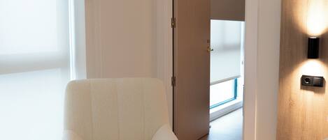 Una habitación moderna con un sillón blanco, una puerta de madera con una lámpara, y vista a un balcón a través de una puerta abierta.