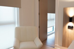 Una habitación moderna con un sillón blanco, una puerta de madera con una lámpara, y vista a un balcón a través de una puerta abierta.