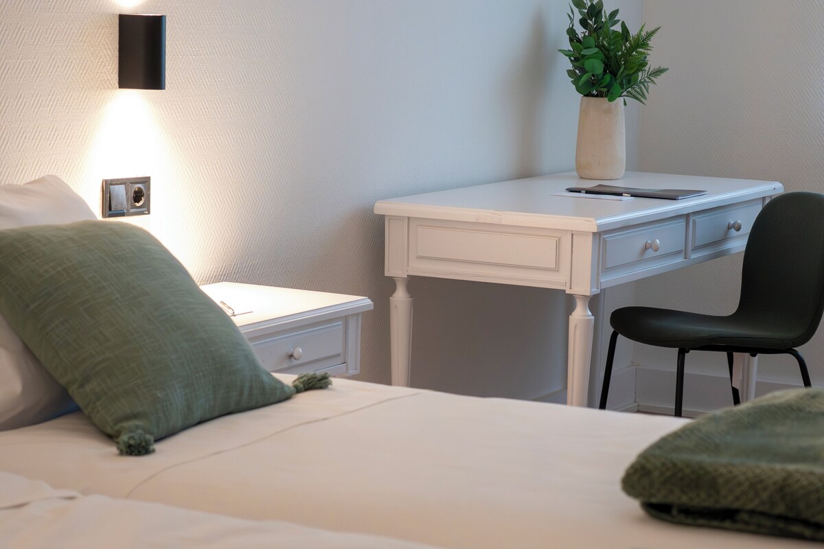 A bedroom with a bed, a desk, a chair, a nightstand, a wall-mounted lamp, and a potted plant.