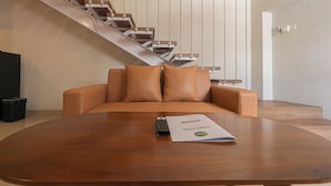 A modern living room with a brown leather sofa, a wooden coffee table, and a remote control on the table. A staircase with wooden steps and metal railings is visible in the background. - Casa Residence Bali (Kerobokan Kaja)