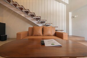 A modern living room with a brown leather sofa, a wooden coffee table, and a remote control on the table. A staircase with wooden steps and metal railings is visible in the background.