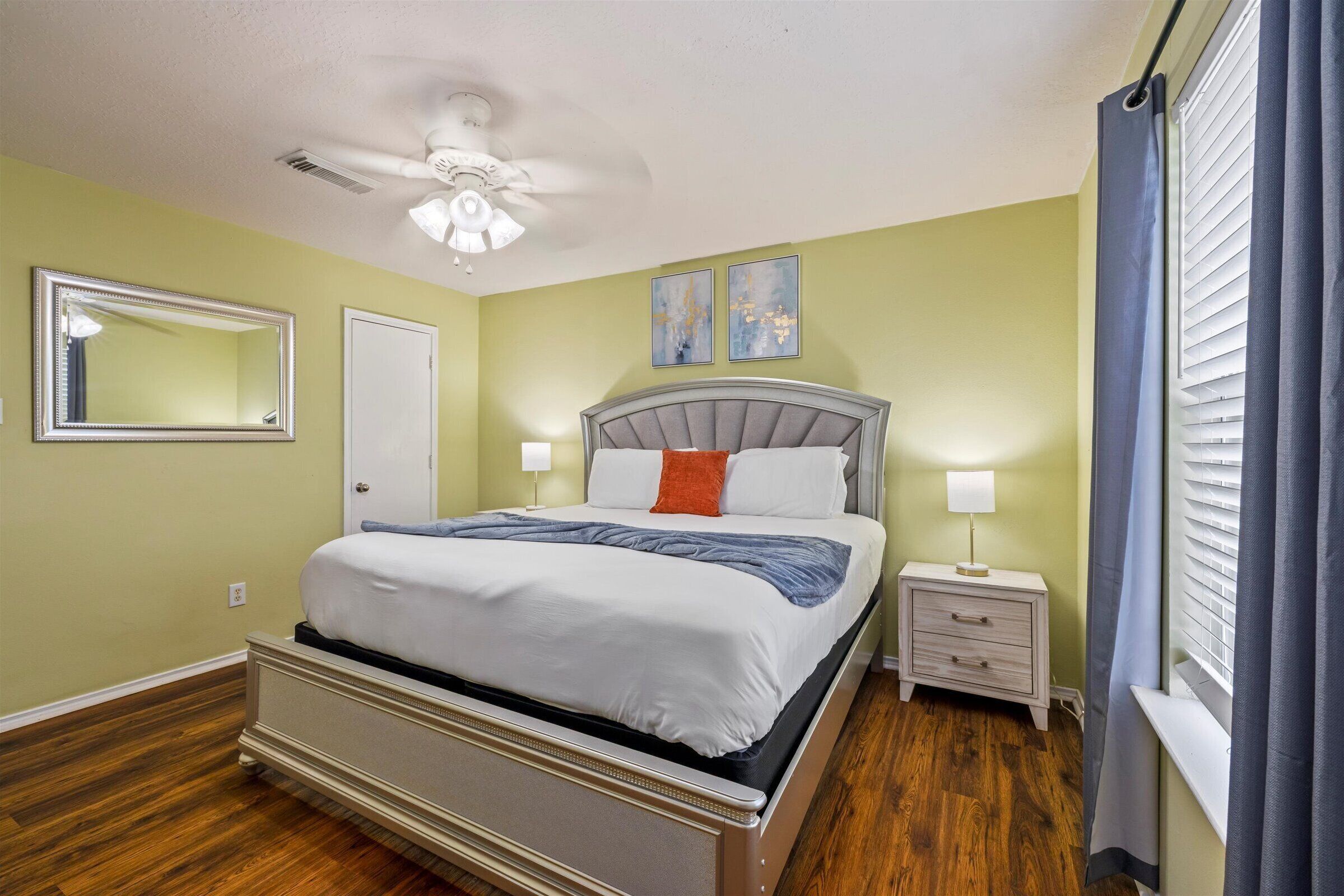 Une chambre à coucher comprenant un lit, une table de chevet, une lampe, un ventilateur de plafond et une œuvre d’art accrochée au mur.