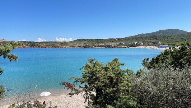 A coastal landscape with a clear blue sea, sandy beach, and a backdrop of hills.