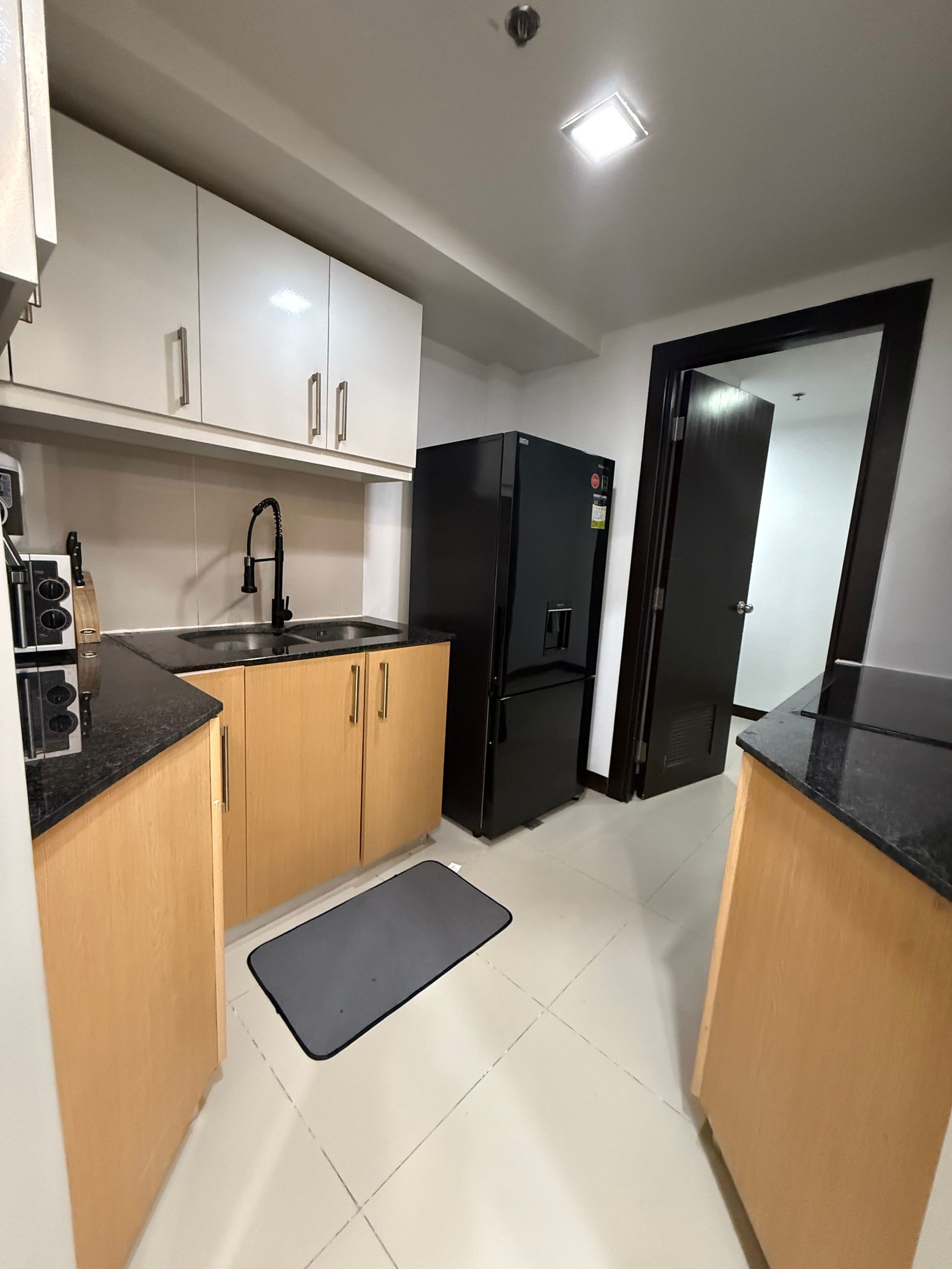 A compact kitchen with white cabinets, a black refrigerator, a sink, and a small mat on the floor.