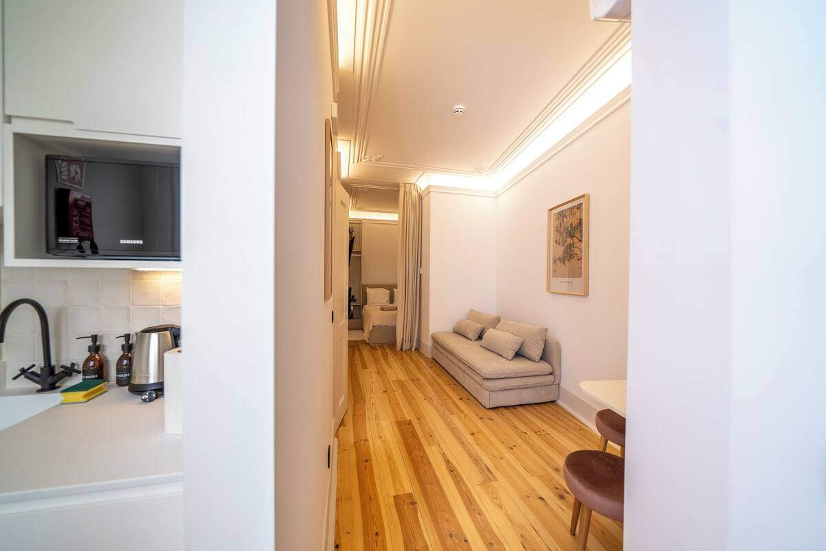 A narrow hallway with a wooden floor leading to a living area with a sofa and a table. A television is mounted on the wall above the sofa.