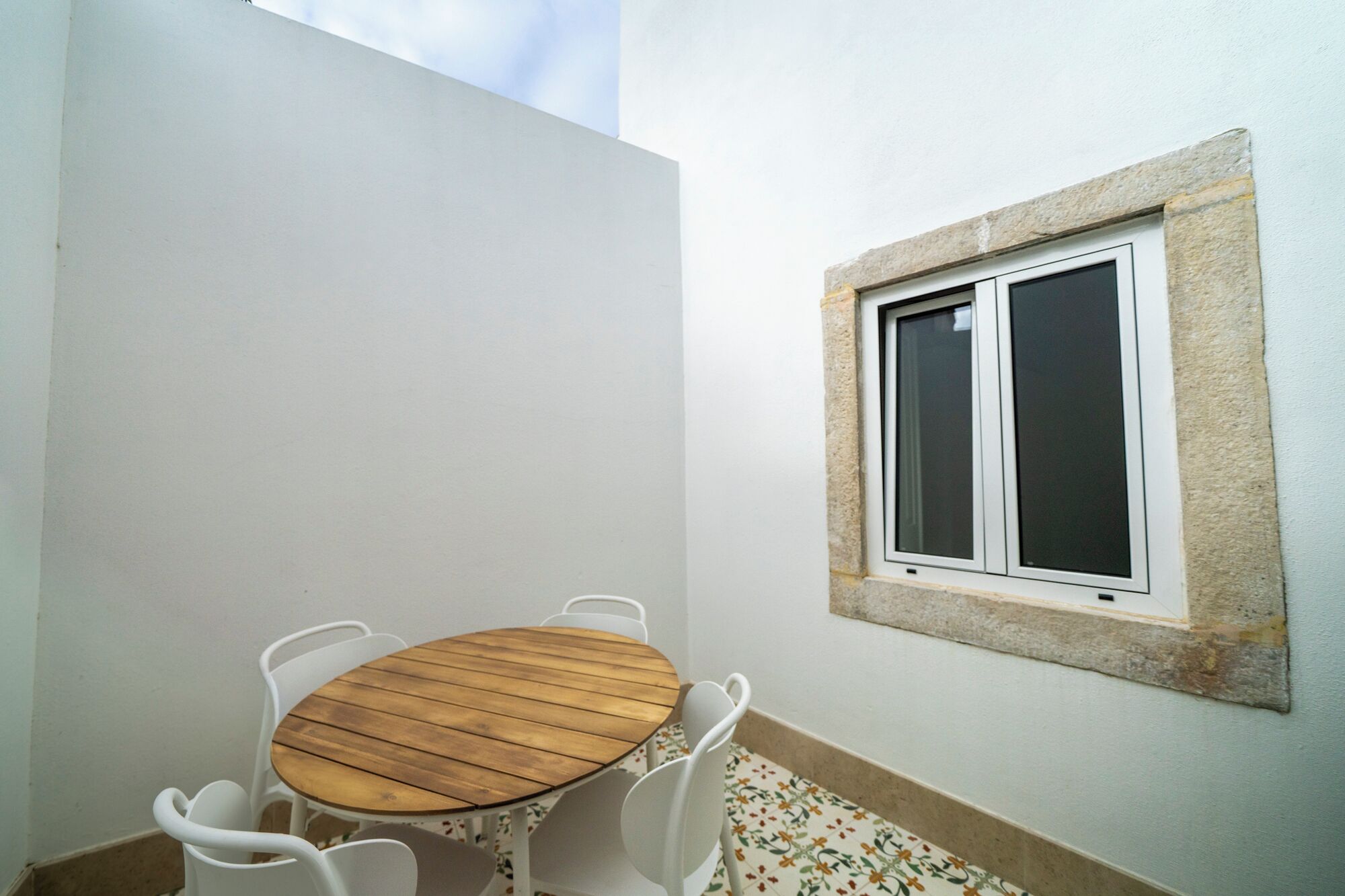 A small room with a round wooden table and white chairs, a window with a stone frame, and a tiled floor with a floral pattern.