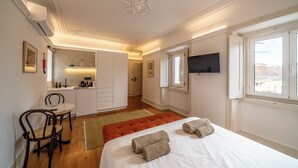 A bedroom with a bed, a TV mounted on the wall, a dining area with chairs, and a kitchen in the background. - Good Old Days (Caldas da Rainha)