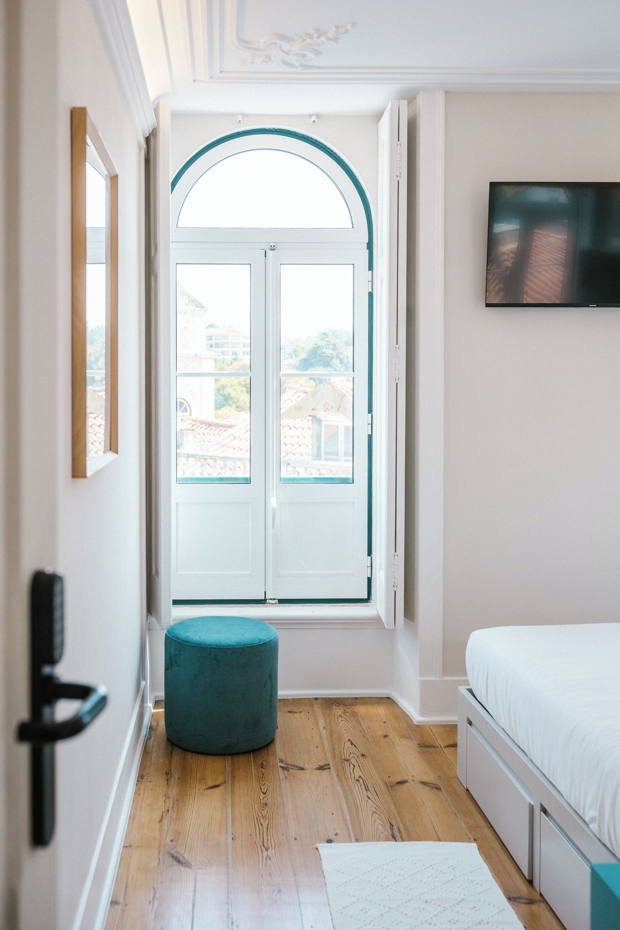 A bedroom with a wooden floor, a white bed, a teal ottoman, a white door with glass panels, and a TV mounted on the wall.