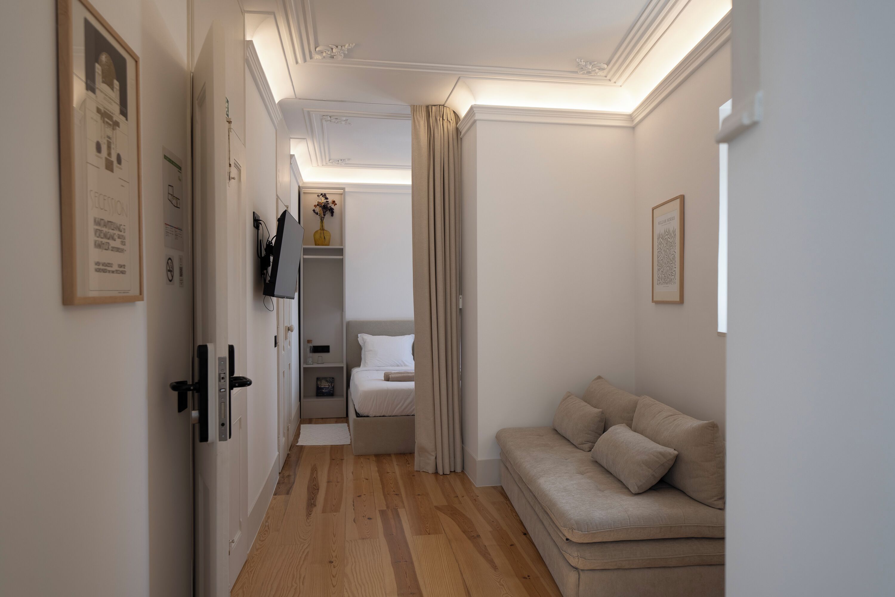 Een smalle gang met een houten vloer die leidt naar een slaapkamer met een bed en nachtkastje.