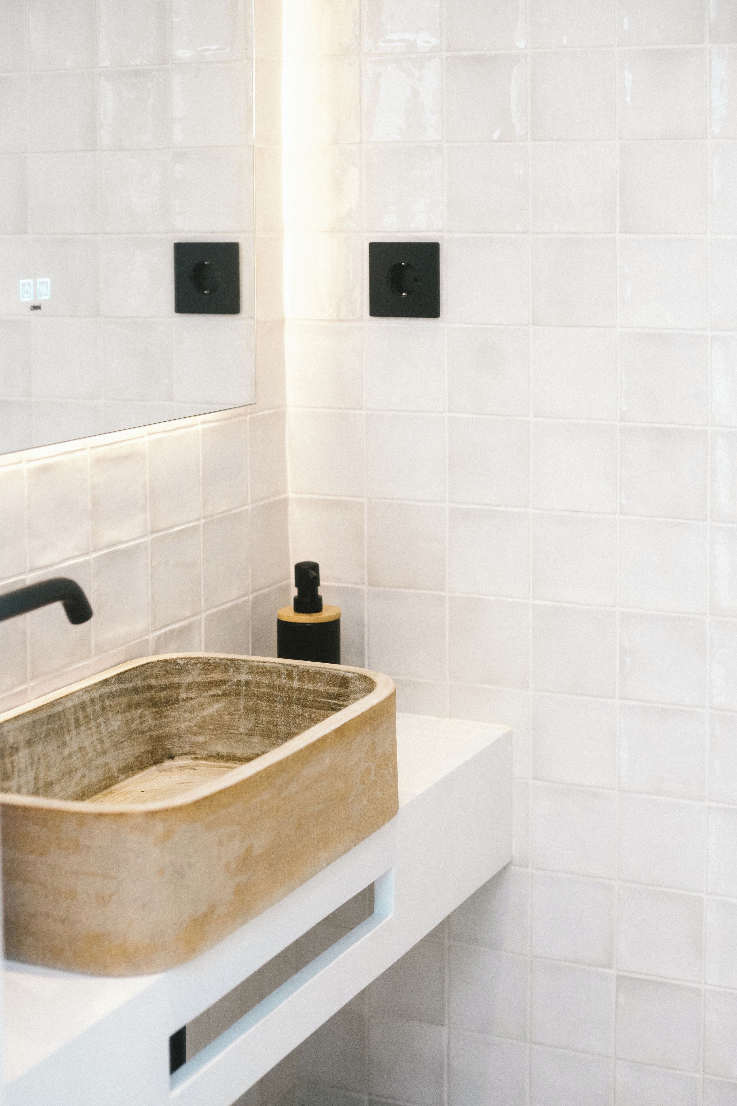 Un baño con un lavabo rectangular, paredes de azulejos blancos y dos interruptores de luz negros fijados a la pared.