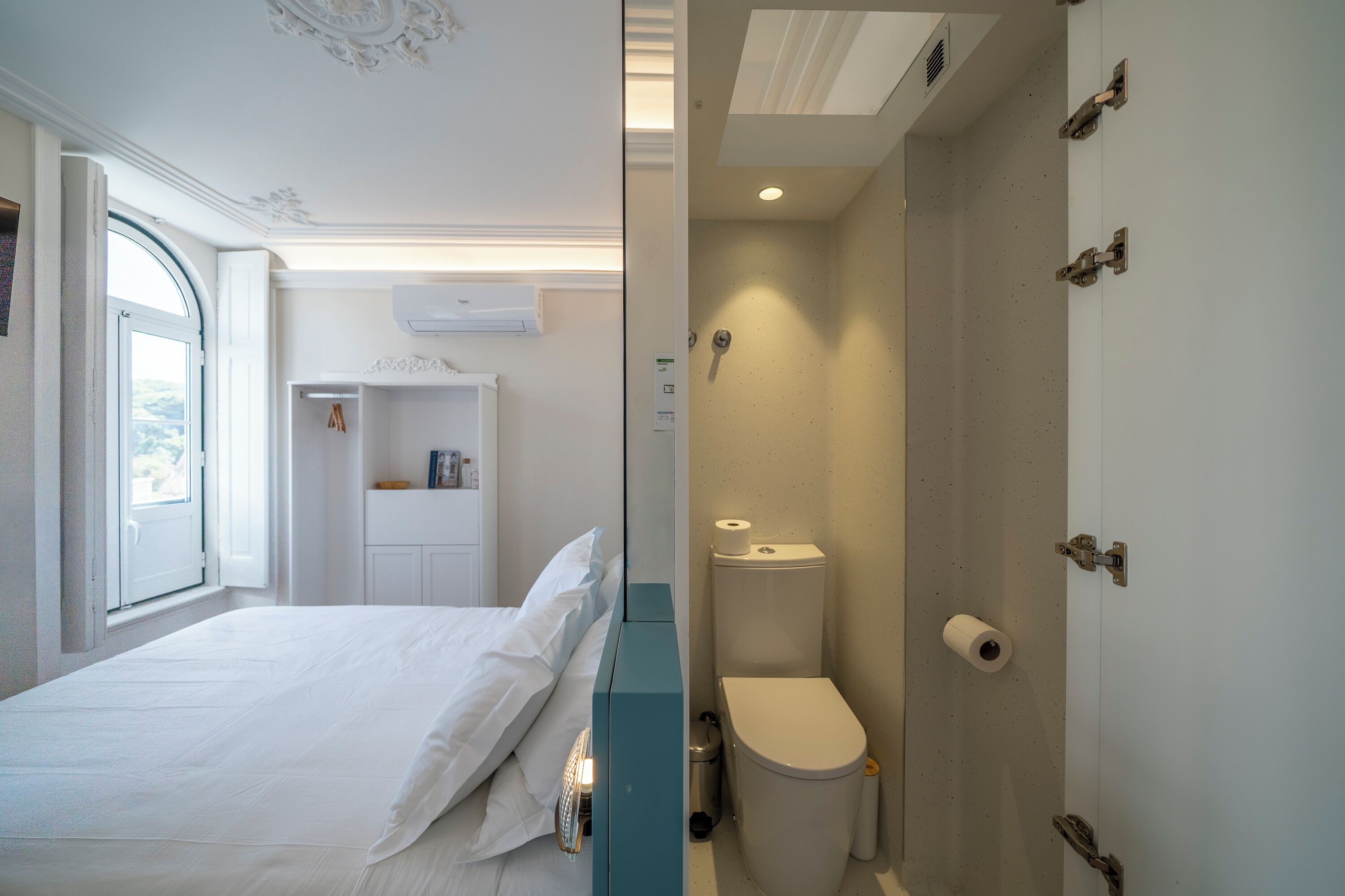 A bedroom with a bed, a window, a wardrobe, and a bathroom with a toilet and shower.