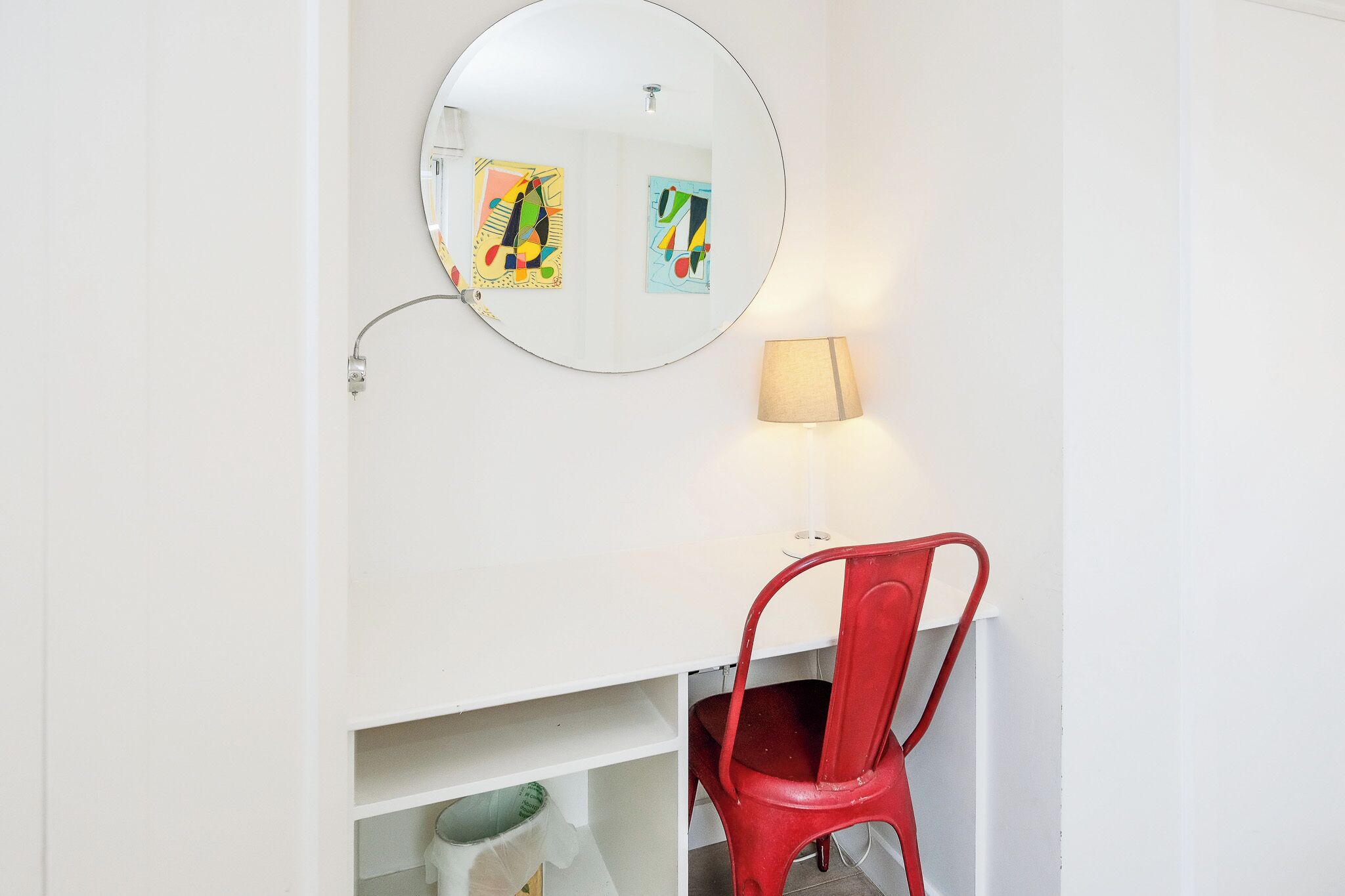 A bathroom with a round mirror, a red chair, and a wall-mounted lamp.