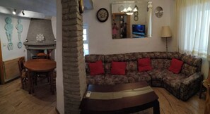 Una sala de estar con chimenea de piedra, un sofá con cojines rojos, una mesa de comedor con sillas y un televisor montado en la pared.