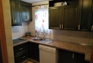Una cocina con armarios de madera oscura, encimera de granito, lavavajillas y ventana con cortina floral.