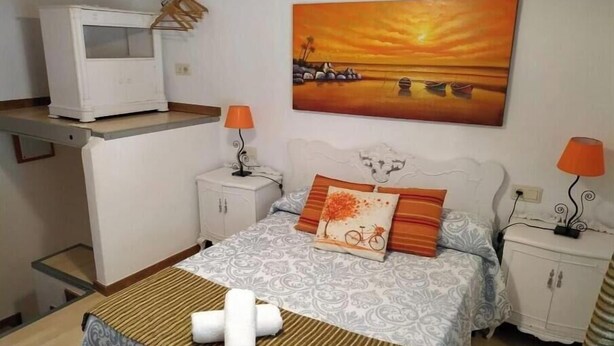Une chambre à coucher comprenant un lit, deux tables de chevet, une commode, un téléviseur et un tableau représentant un coucher de soleil sur l’eau.